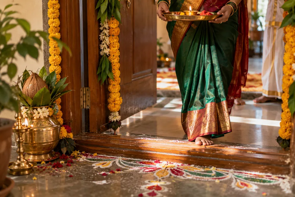A woman in a green silk saree carrying a lit lamp across a decorated doorway with marigold garlands and rangoli